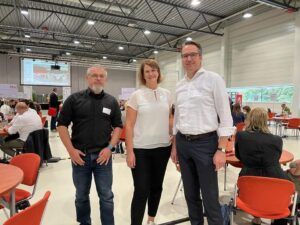 Deutscher Wohnbau Verbund auf der baupressekompakt: Alexander Schroeder, Jacqueline Paulic und Bert Große Drei Personen stehen in einer Veranstaltungshalle für ein Foto: Alexander Schroeder, Jacqueline Paulic und Bert Große.