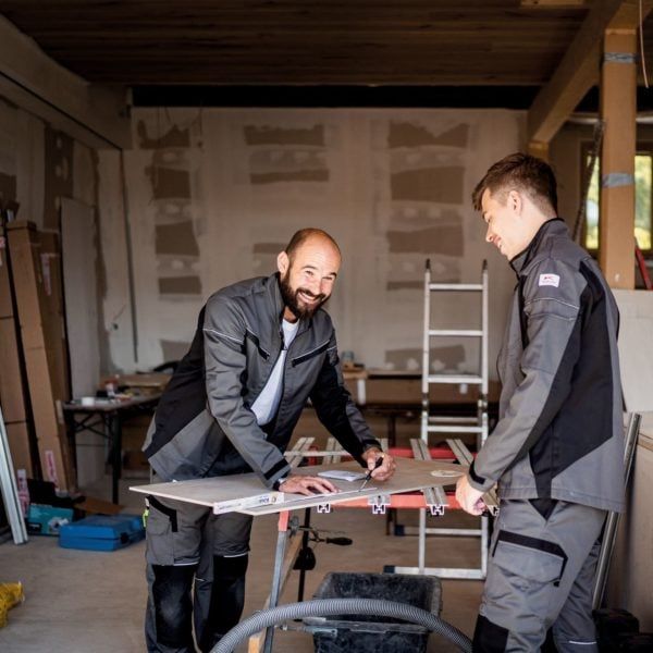 Zwei Handwerker planen Zuschnitt in Werkstatt Zwei Handwerker in Arbeitskleidung stehen in einer Werkstatt an einer Werkbank und messen eine Platte aus.