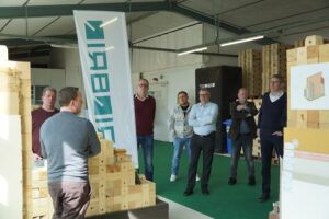 Vorführung an Holzbausystem-Modell in der Werkhalle Personen stehen vor einem aufgebauten Holzmodul-Modell und hören einer Erklärung zu.