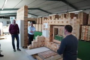 Besuchergruppe in Werkhalle mit modularen Holzbausteinen Mehrere Personen stehen in einer Werkhalle zwischen hohen Stapeln modularer Holzbausteine.