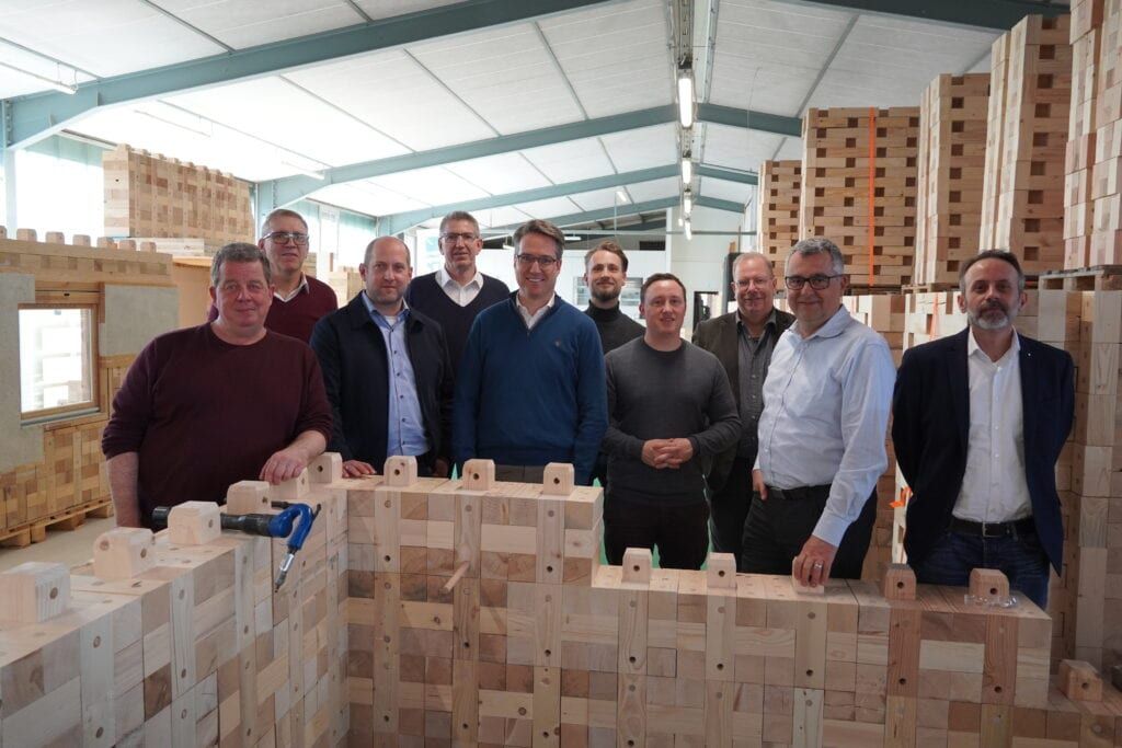 Besuchergruppe in Holzbau-Werkhalle vor modularen Holzbausteinen Besuchergruppe steht in einer Werkhalle zwischen großen Stapeln modularer Holzbausteine.