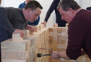 Montagearbeit an modularen Holzbausteinen Zwei Personen arbeiten nah am Holzbaustein-Aufbau; eine Person hält ein Werkzeug an der Konstruktion.