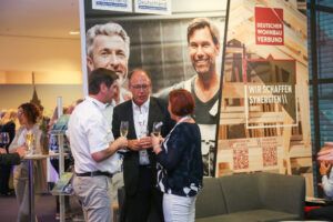Gespräch am Messestand in einer Ausstellungshalle Mehrere Personen stehen an einem Messestand im Gespräch; im Hintergrund sind Werbebanner mit Porträtfotos zu sehen.