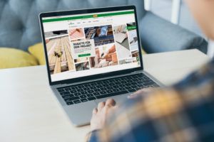 Online-Baustoffhandel am Laptop Person sitzt vor einem Laptop; auf dem Bildschirm ist eine Website mit Produktkacheln zu sehen.