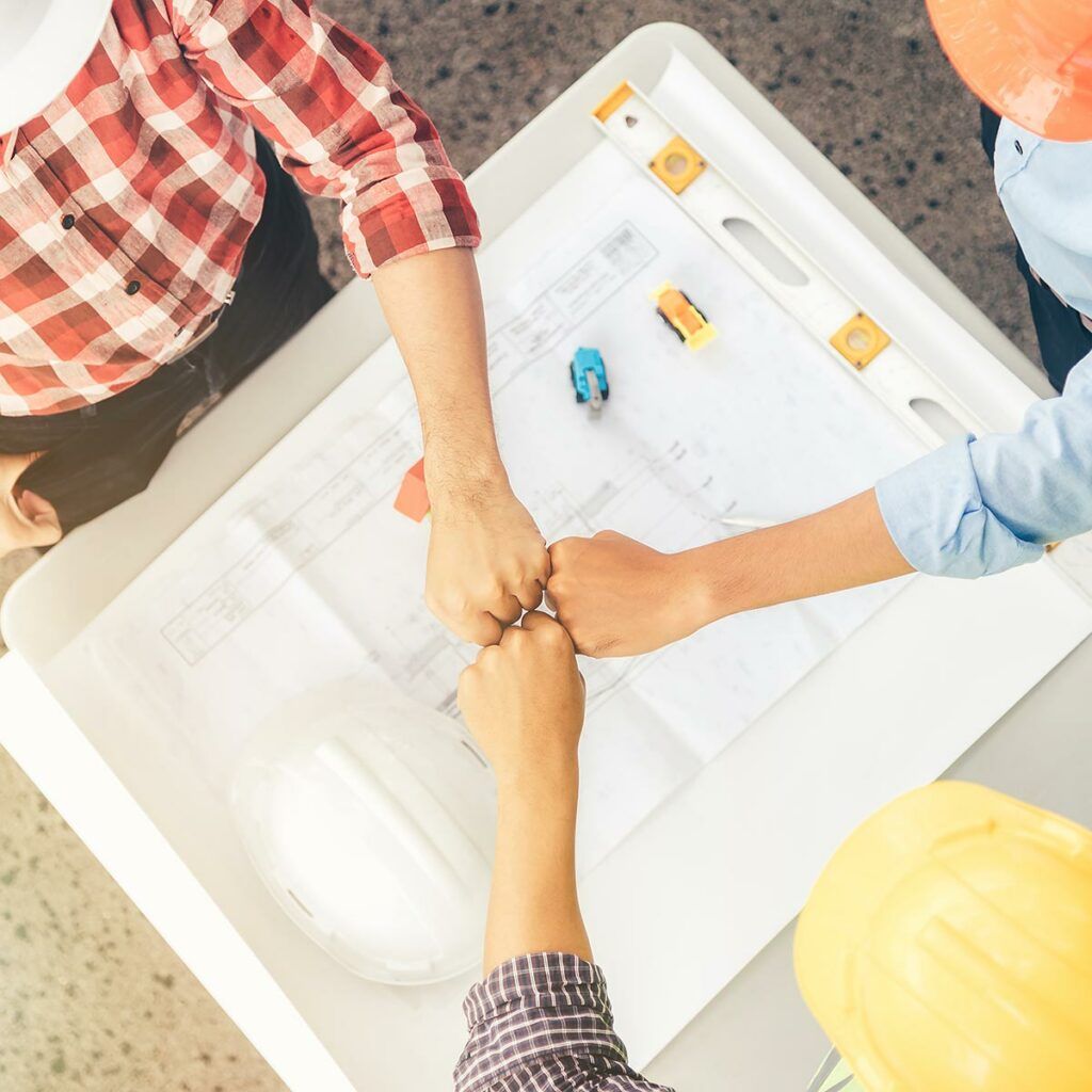 Teamwork am Bauplan Drei Personen stoßen mit den Fäusten über einem Bauplan zusammen; Bauhelme liegen auf dem Tisch.