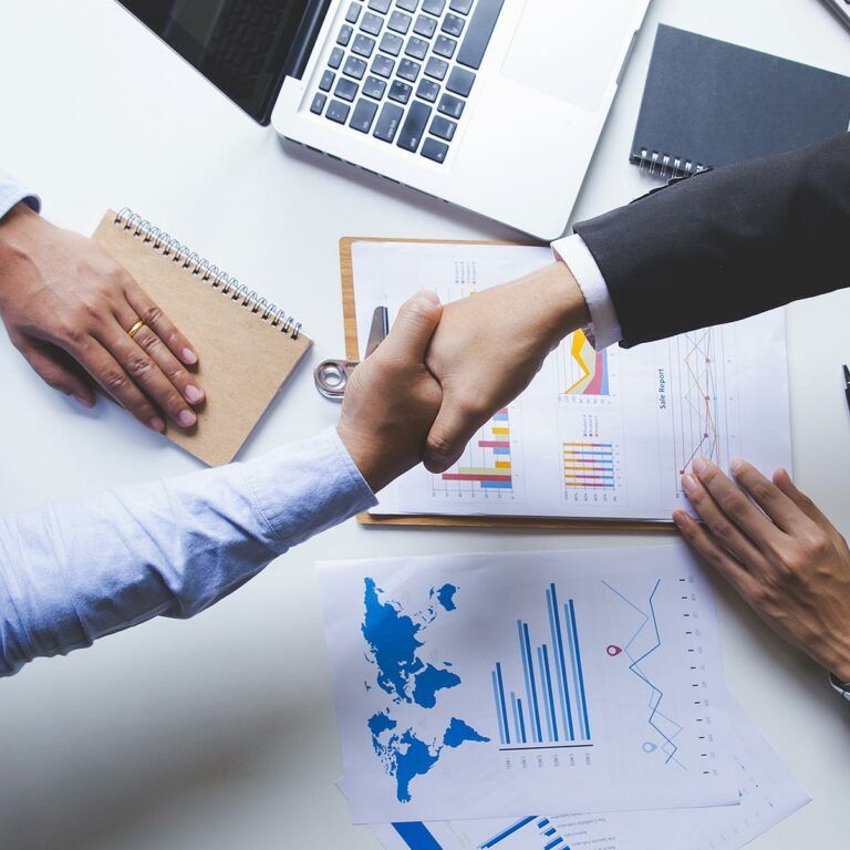 Zwei Personen geben sich die Hand über Unterlagen mit Diagrammen auf einem Schreibtisch.