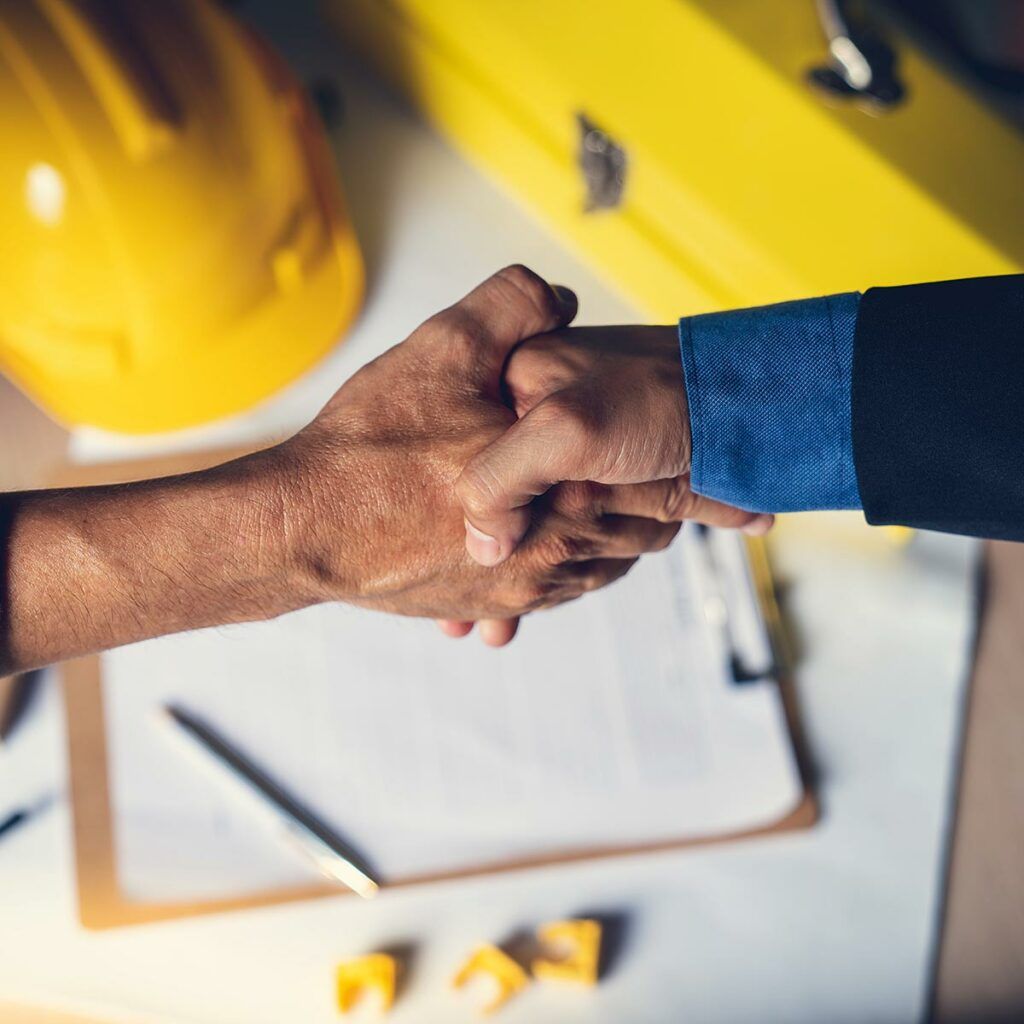 Handschlag auf Baustelle Zwei Personen geben sich die Hand; im Hintergrund liegen Bauhelm und Planunterlagen.