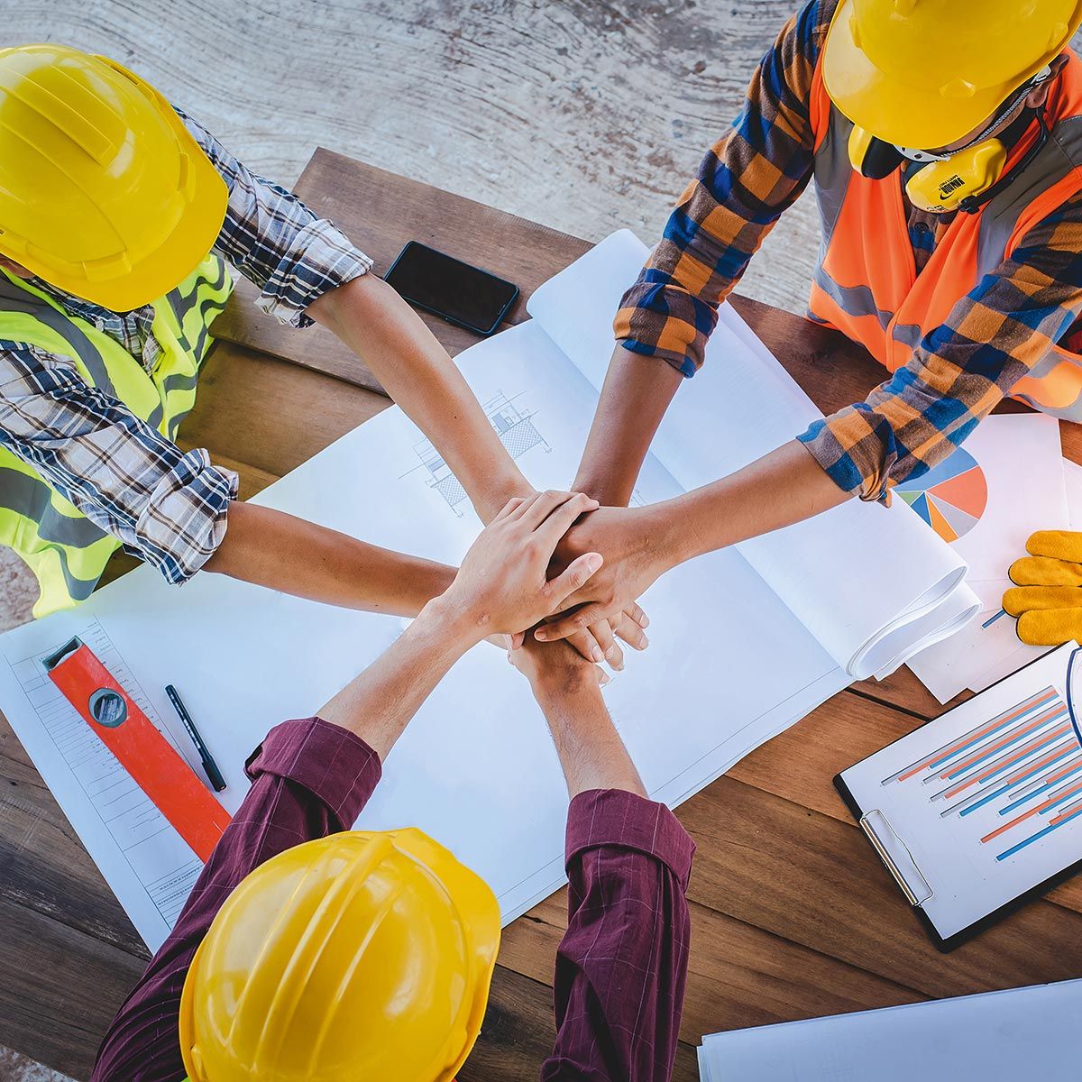 Teamwork auf der Baustelle Drei Personen mit Bauhelmen legen die Hände übereinander über Bauplänen auf einem Tisch.
